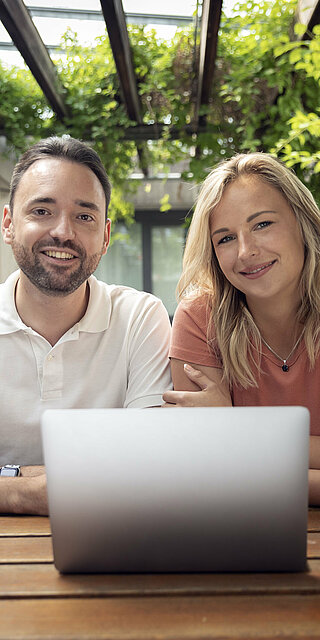 A man and a woman sit in front of a laptop in a courtyard