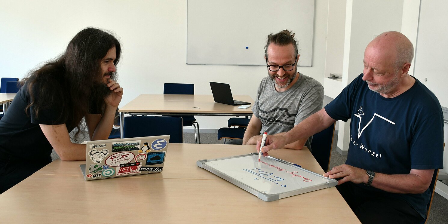 Drei Männer sitzen an einem Tisch, einer schreibt auf eine Tafel.