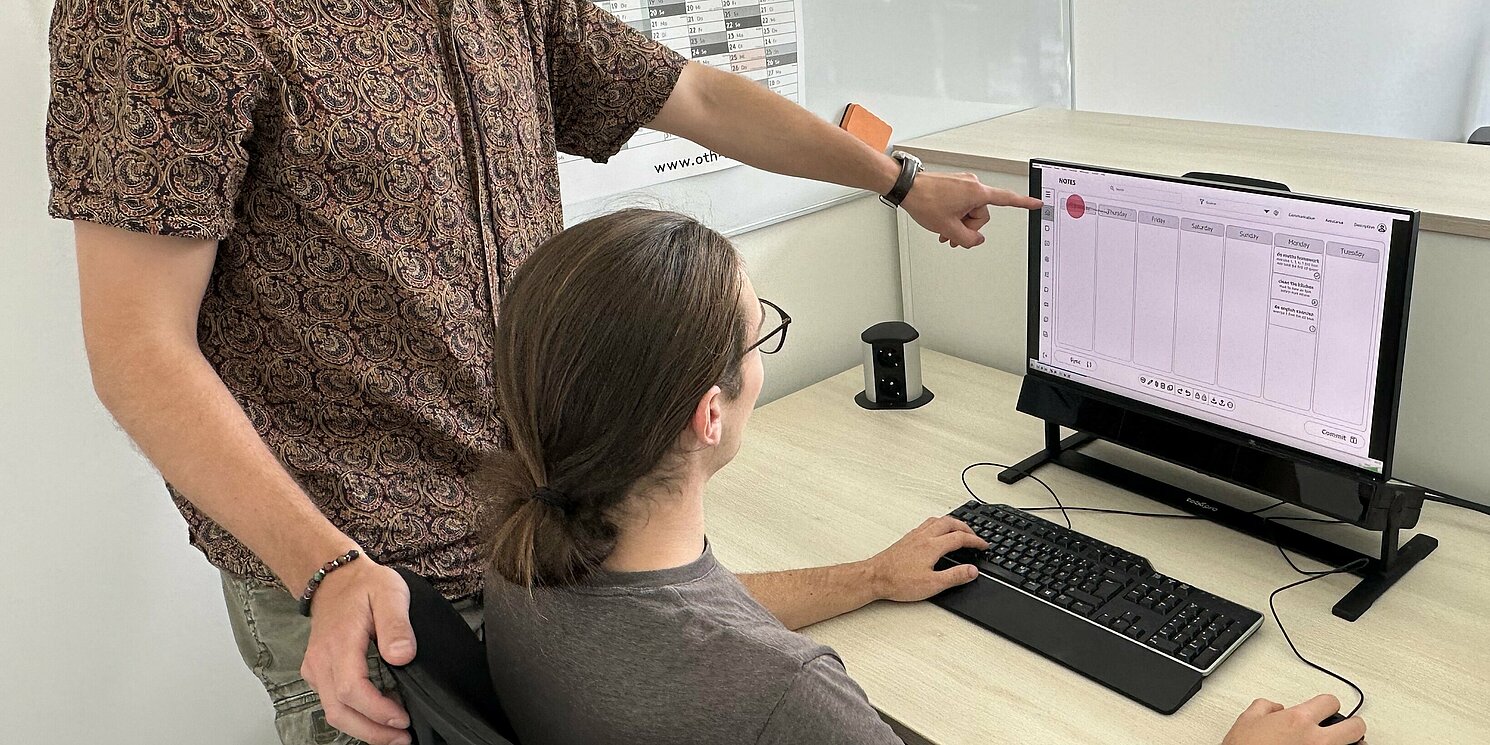 Ein Mann sitzt vor einem PC, ein weiterer Mann steht daneben und zeigt auf den Bildschirm.